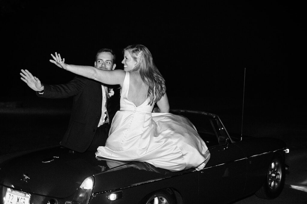 Bride and groom waving goodbye to guests while riding away in their getaway car.