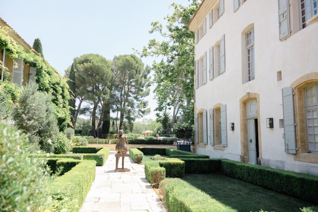 A stone path framed by boxwood hedges, with a bronze statue in the center, between vine-covered walls and a white house with blue shutters